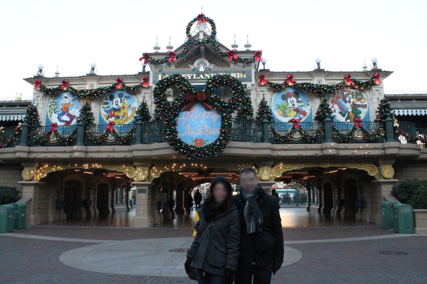 Los dos, con abrigo y bufanda, ante la entrada decorada de Disneyland Paris® al atardecer, con la explanada casi vacía, en diciembre de 2013